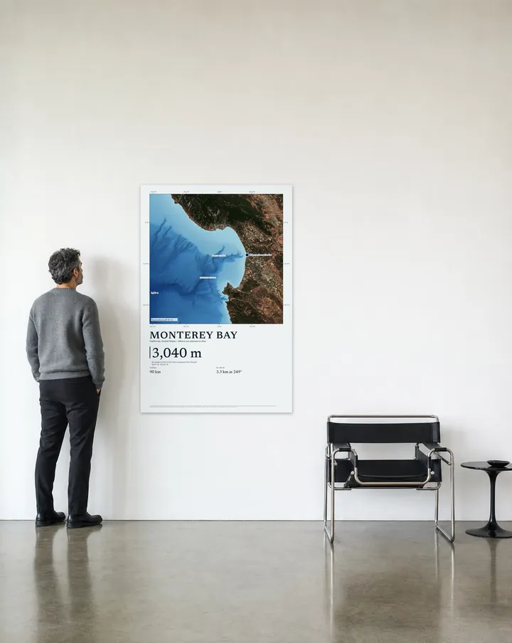 Generated scene: a loft, beside a lounge chair. On the wall, our real Satellite drawing of Monterey Bay at 40 × 60 in, set at true scale beside a man 6 ft tall.