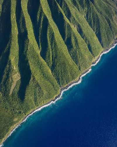 Generated scene: a volcanic island's green ridges from high above, the sea turquoise over the shelf and navy past it.