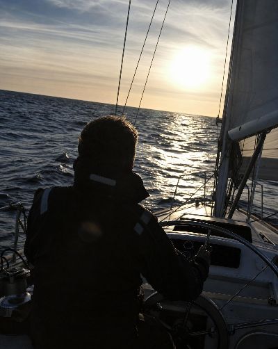 Generated scene: a sailor at the wheel of a small yacht on open water in late sun.